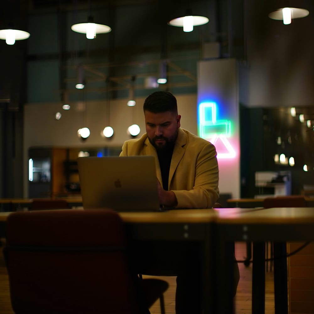 Laptop-Nutzer in einem modernen, urbanen Working Space bei Nacht, beleuchtet von bunten Neonlichtern, fokussiert auf seinem Gerät für effizientes Arbeiten in einer kreativen Umgebung.