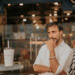 Bergsamer Mann in einem Café mit nachdenklicher Pose, sitzend an einem Tisch mit einem großen Getränk, klimatisiert, modernes Ambiente, entspannte Atmosphäre, Eventualität eines Gesprächs.